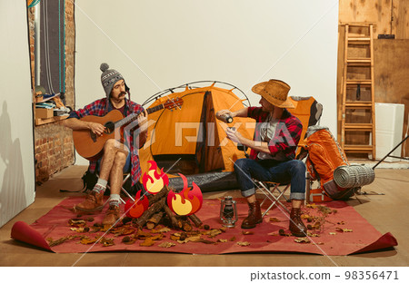 Playing guitar, drinking tea. Two men imagining, recreating camping activity indoors with necessary equipment. Concept of quarantine, travelling, imagination 98356471