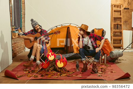 Two men imagining, recreating camping activity indoors with necessary equipment. Playing guitar. Concept of quarantine, travelling, imagination Two men imagining, recreating camping activity indoors with necessary equipment. Playing guitar. Concept of quarantine, travelling, imagination 98356472