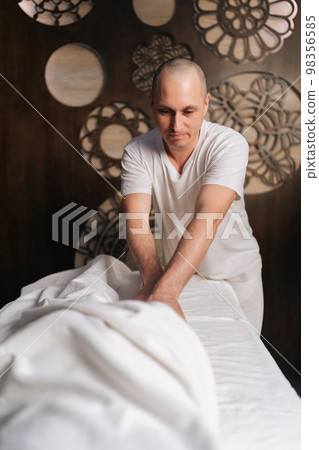 Vertical portrait of bald male masseur performing anti-cellulite massage on buttocks of relaxed female lying on table in spa salon. Beautiful young woman getting relaxing lymphatic drainage massage. 98356585