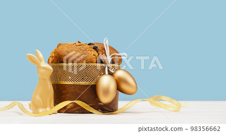 Happy Easter banner. Bunny and Italian Easter Decorated Panettone and golden eggs on white table and festive ribbon. Wide banner on blue. Traditional bakery. Christianity 98356662