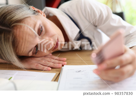 Tired female doctor laying on her work desk and looking at smartphone screen. Tired female doctor laying on her work desk and looking at smartphone screen. 98357830