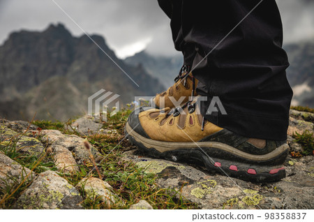 Close-up of legs in trekking boots against the backdrop of an alpine mountain range in a valley 98358837