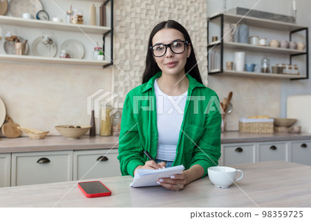 Portrait of a young woman teacher in glasses sitting at home in the kitchen in front of the camera. Works from home online. He is holding a notebook with a pen in his hands, smiling. 98359725