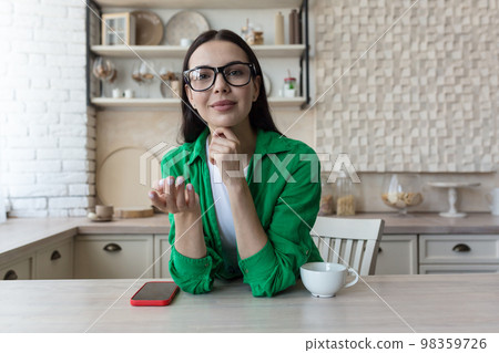 A young beautiful female blogger is sitting at home in the kitchen and talking online to the camera, explaining with her hands, smiling. A young beautiful female blogger is sitting at home in the kitchen and talking online to the camera, explaining with her hands, smiling. 98359726