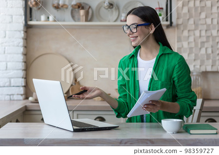 A young female student, a freelancer studies, works online at home from a laptop. He sits at the table, listens to the lecture, holds a notebook with a pen in his hands, writes down, explains. 98359727