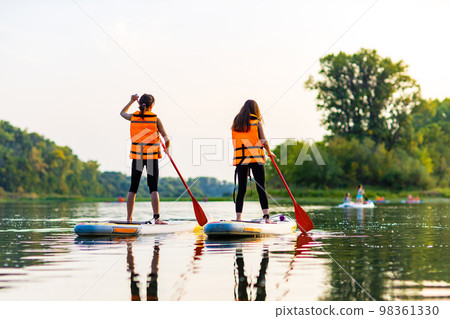 two young women in orange life jacket on supboard at river 98361330