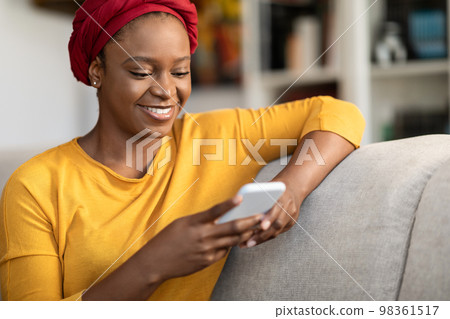 Positive relaxed black woman sitting on couch, using smartphone Positive relaxed black woman sitting on couch, using smartphone 98361517