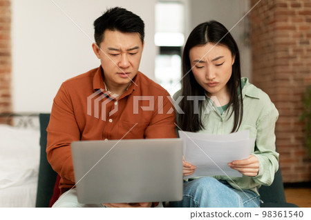 Upset korean spouses checking bills, reading documents while sitting on couch and using laptop. Family budget concept 98361540