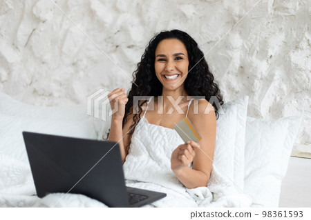 Glad arab millennial curly lady woke up, sitting on white bed with laptop, making victory and success gesture 98361593