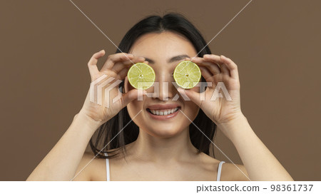 Vitamins for beauty and skin care. Asian lady covering eyes with lime halves, standing over brown background, panorama 98361737