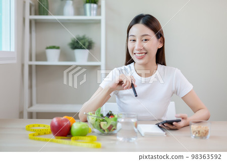 Females using smartphones taking notes eating healthy food each day with vegetables fruit on table. Females using smartphones taking notes eating healthy food each day with vegetables fruit on table. 98363592