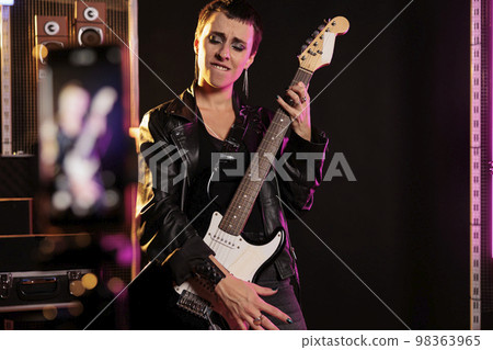 Rebel rockstar holding electric guitar playing heavy metal song recording performance with modern phone during music session in audio studio. Woman performer working at new grunge album before tour 98363965