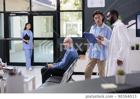 African american doctor explaining disease expertise to asian patient during checkup visit consultation in hospital waiting area. Young man filling medical insurance documents, medicine service 98364013