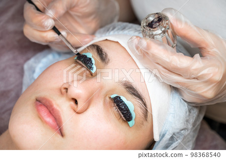 Close-up portrait of a woman on eyelash lamination procedure. The master applies tint to the eyelashes.  98368540