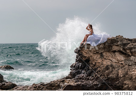 A woman in a storm sits on a stone in the sea. Dressed in a white long dress, waves crash against the rocks and white spray rises above her. 98375812