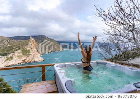 Take time for yourself. Outdoor swimsuit with mountain and sea views. A woman in a black swimsuit is relaxing in the hotel pool, admiring the view Take time for yourself. Outdoor swimsuit with mountain and sea views. A woman in a black swimsuit is relaxing in the hotel pool, admiring the view 98375870