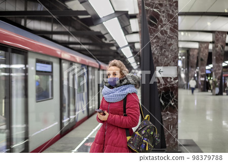 A young woman in a medical mask and a vivid magenta coat with a scarf holding a phone in her hand, waiting for a train in the subway 98379788