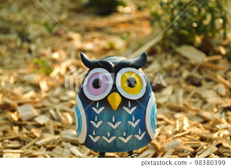 A toy of a surprised blue owl with multi-colored eyes close-up stands on wooden sawdust in the light of the evening sun A toy of a surprised blue owl with multi-colored eyes close-up stands on wooden sawdust in the light of the evening sun 98380399
