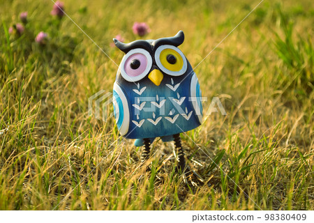 A toy of a surprised blue-colored owl with multi-colored eyes close-up stands in the middle of the grass in the light of the evening sun 98380409