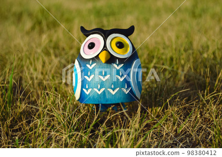 A toy of a surprised blue-colored owl with multi-colored eyes close-up stands in the middle of the grass in the light of the evening sun 98380412