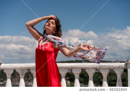 A woman in a red dress against a blue sky and white clouds with a developing scarf around her neck. She turned her face to the wind and posed for the camera. 98382804