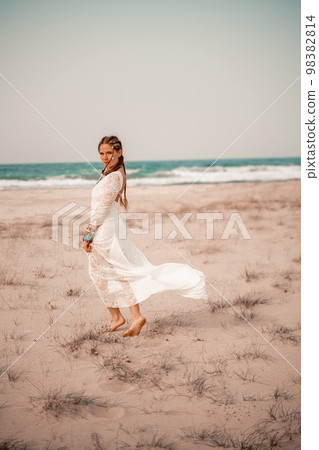 Model in boho style in a white long dress and silver jewelry on the beach. Her hair is braided, and there are many bracelets on her arms. 98382814