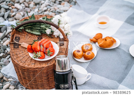 food, holidays and celebration concept - close picnic basket with thermos with tea, rolls, strawberries, flowers on the beach 98389625