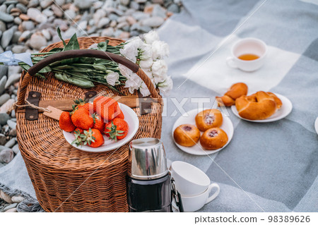 food, holidays and celebration concept - close picnic basket with thermos with tea, rolls, strawberries, flowers on the beach 98389626