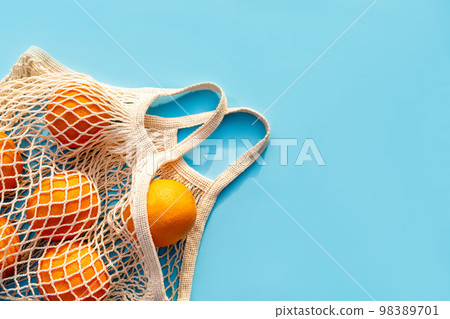 Mesh white cotton reusable bag with oranges on a blue background. Environmentally friendly shopping, the environment and the concept of healthy foods, citrus fruits with vitamin C. Place for text 98389701