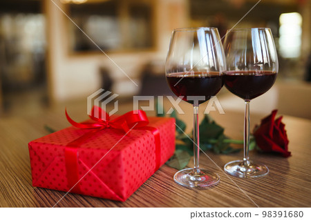 Valentine's day celebration concept. Two glasses of wine, red rose red gift box on wooden table. Valentine's day celebration concept. Two glasses of wine, red rose red gift box on wooden table. 98391680