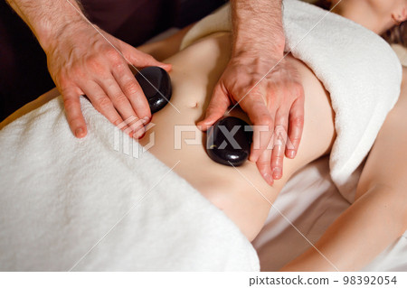 Close up view of man hands puts warm massage stones on the female patient's stomach 98392054
