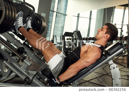 Young man is engaged on a modern simulator in the gym. Male athlete is doing exercises for legs with training machine. Young man is engaged on a modern simulator in the gym. Male athlete is doing exercises for legs with training machine. 98392256