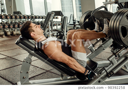 Man Using A Press Machine In A Fitness Club. Strong man doing an exercise on its feet in the simulator. Side view. 98392258