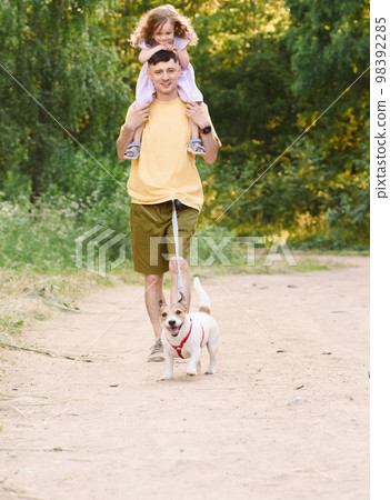 Father and daughter walking outside by countryside dirt road with pet dog on waist leash 98392285
