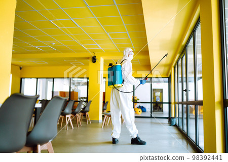 Contractor disinfecting office for COVID-19 coronavirus. A disinfector in a protective suit and mask Contractor disinfecting office for COVID-19 coronavirus. A disinfector in a protective suit and mask 98392441