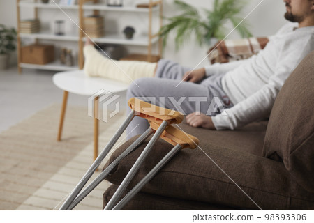 Close up of crutches leaned on the sofa, and a man with a broken leg sitting in the background 98393306
