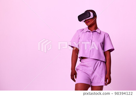 Young african american positive woman in black virtual reality glasses with vr headset posing, looking, playing and touching hands on purple background. 98394122