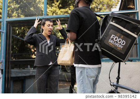 Angry customer shouting at food delivery service courier, problem with takeaway meal order. African american deliveryman giving impulsive client lunch package in front of company building Angry customer shouting at food delivery service courier, problem with takeaway meal order. African american deliveryman giving impulsive client lunch package in front of company building 98395142