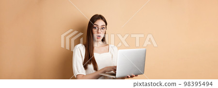 Surprised girl using laptop and gasping amazed, say wow, looking in glasses at camera, beige background 98395494