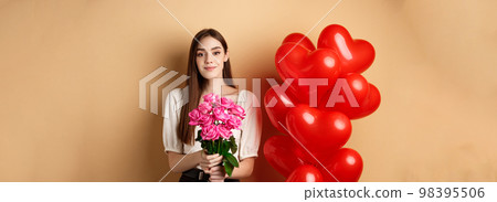 Beautiful girl holding bouquet of pink roses and smiling at camera, going on romantic date, standing near valentines heart balloons, beige background 98395506