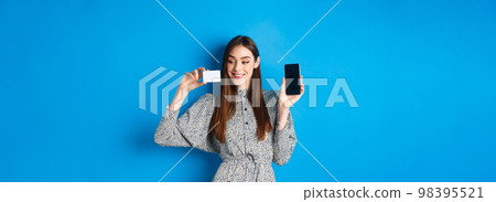 Online shopping. Beautiful female model showing empty cellphone screen and plastic credit card, smiling pleased, buying in internet store, blue background 98395521