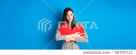 Valentines day. Romantinc girl in dress hugging big red heart cutout, close eyes and smile with dreamy face, imaging sensual date, standing on blue background 98395522