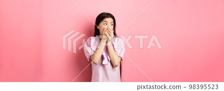Shocked and startled asian woman looking aside at logo, covering mouth with hands speechless, standing in dress on pink background 98395523