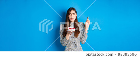 Hopeful birthday girl making wish on cake, cross fingers for good luck, standing in trendy dress against blue background Hopeful birthday girl making wish on cake, cross fingers for good luck, standing in trendy dress against blue background 98395590