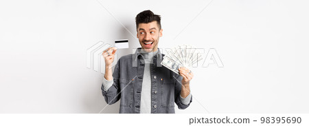 Excited guy holding plastic credit card and dollar bills, standing amused on white background 98395690