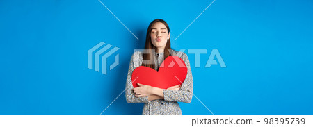 Valentines day. Romantic beautiful woman close eyes and pucker lips for kiss, holding big red heart cutout, kissing you, standing on blue background 98395739