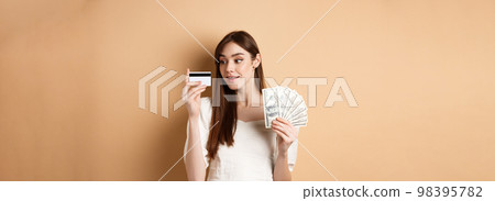 Dreamy girl looking at plastic credit card and thinking of shopping, holding dollar bills, standing on beige background Dreamy girl looking at plastic credit card and thinking of shopping, holding dollar bills, standing on beige background 98395782