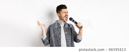 Handsome guy singing in mic with passion, perform for audience, holding microphone and gesturing, standing on white background Handsome guy singing in mic with passion, perform for audience, holding microphone and gesturing, standing on white background 98395813