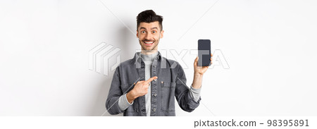 Handsome smiling man pointing finger at smartphone screen, showing mobile phone display with happy face, standing against white background Handsome smiling man pointing finger at smartphone screen, showing mobile phone display with happy face, standing against white background 98395891