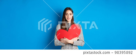 Valentines day. Attractive young woman searching for love, holding big red heart cutout and smiling at camera, standing in dress on blue background Valentines day. Attractive young woman searching for love, holding big red heart cutout and smiling at camera, standing in dress on blue background 98395892